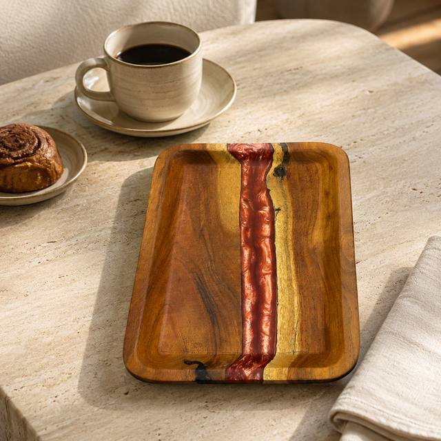 A rectangular wooden serving tray with a polished finish and a central red and yellow wood grain accent, displayed on a light stone table next to a cup of coffee, a pastry, and a folded napkin.